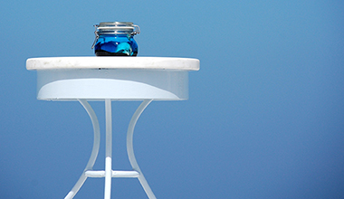 Small glass bowl with blue liquid on a white metal table against a blue background.
